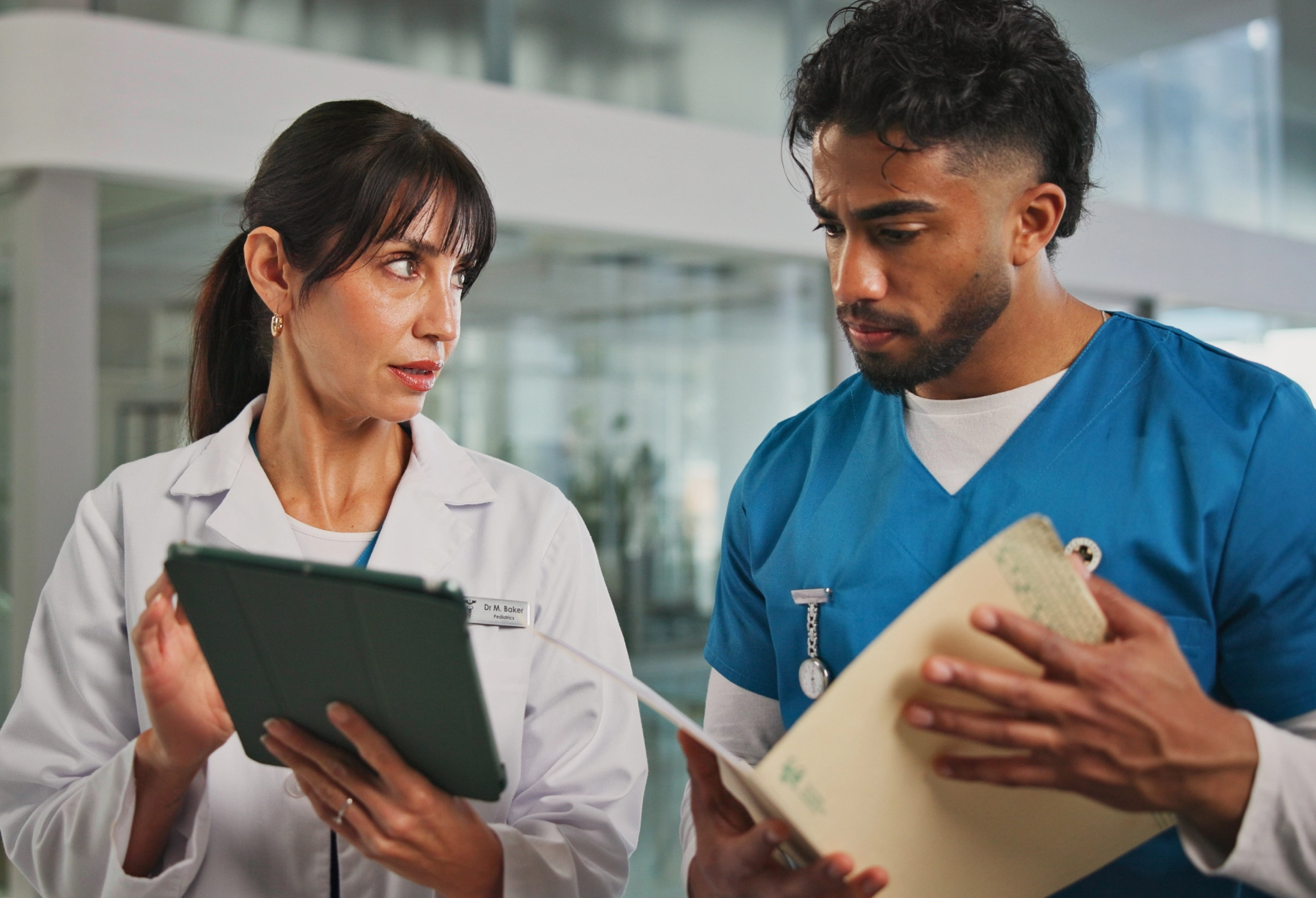 Two healthcare professionals, one in a white coat and another in blue scrubs, standing while looking at a tablet